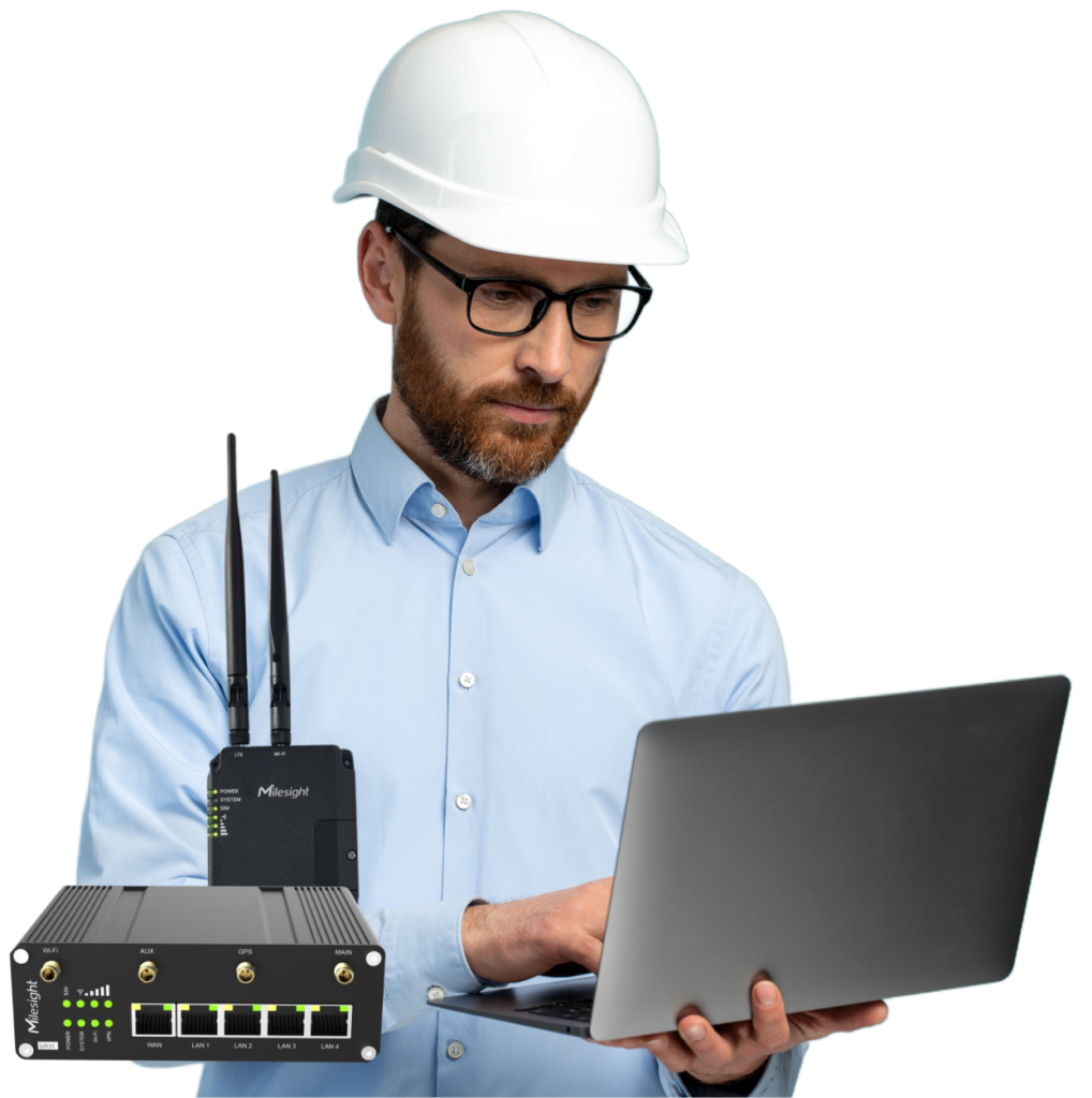 A construction worker wearing a white hard hat and glasses, working on a laptop with industrial networking equipment displayed.