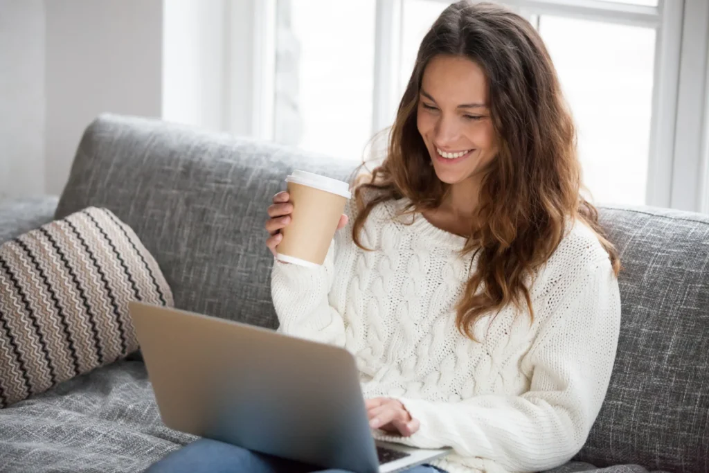 Smiling young woman enjoying morning coffee using laptop sitting at home on sofa, attractive happy girl typing on computer having fun online or chatting with friends while relaxing on couch